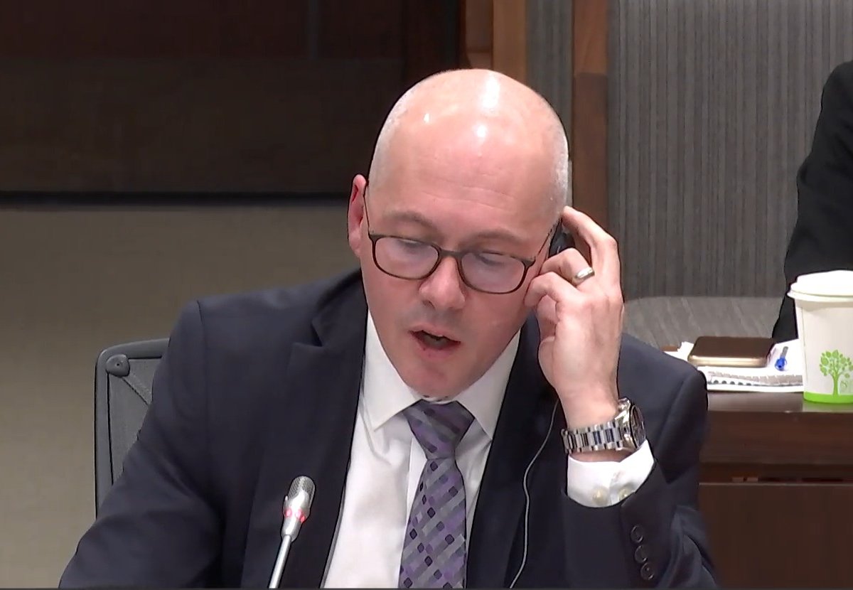 Cameron MacDonald at the committee table, hand at his earpiece